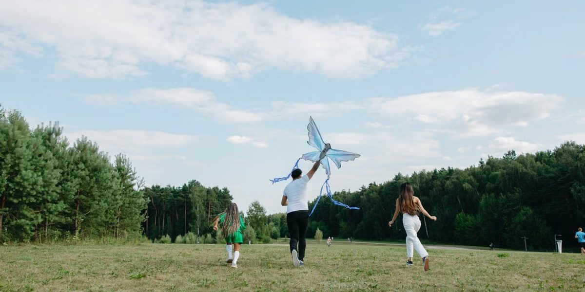 A family runs joyfully while flying a kite in a sunny park setting.
