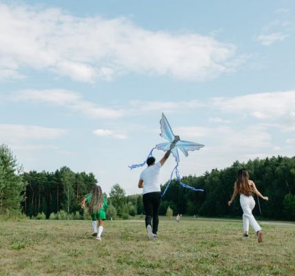 A family runs joyfully while flying a kite in a sunny park setting.